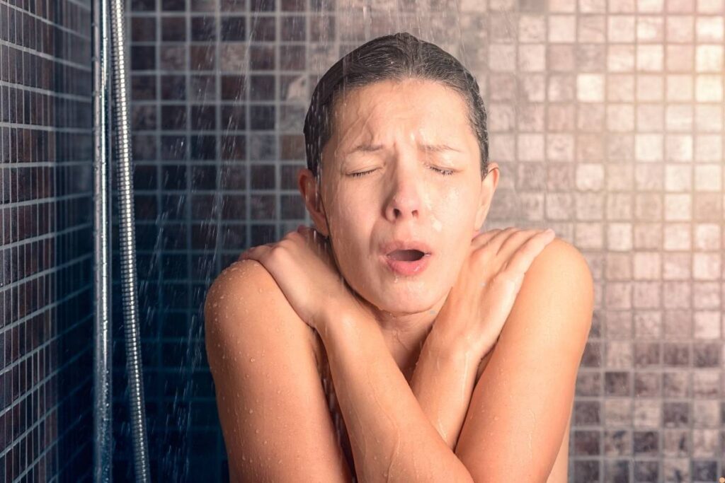 Woman reacting to cold water in a tiled shower, arms crossed tightly for warmth, showing discomfort from a short hot water supply in a residential bathroom—relating to hot water repair in Albury homes.