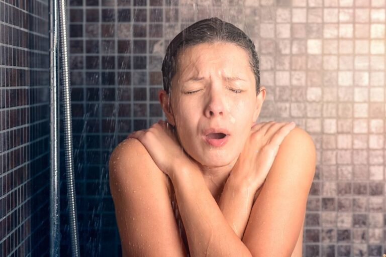 Woman reacting to cold water in a tiled shower, arms crossed tightly for warmth, showing discomfort from a short hot water supply in a residential bathroom—relating to hot water repair in Albury homes.