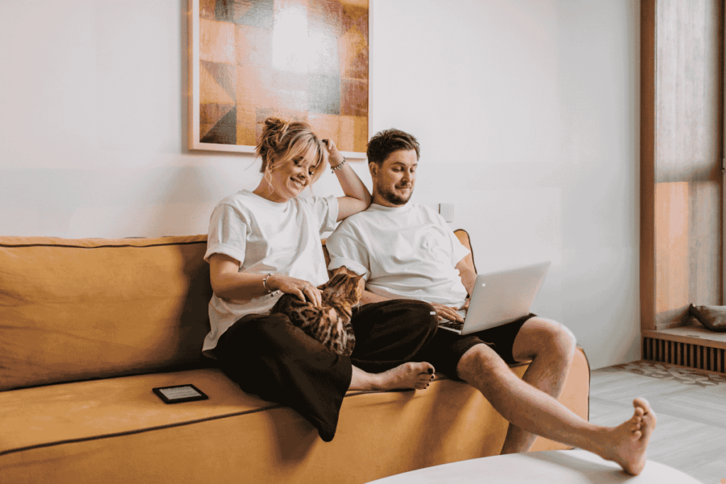 A couple sitting on a tan sofa at home in Albury Wodonga, smiling and relaxed with a cat on their lap, enjoying cool indoor comfort from an evaporative air conditioner on a warm summer day.