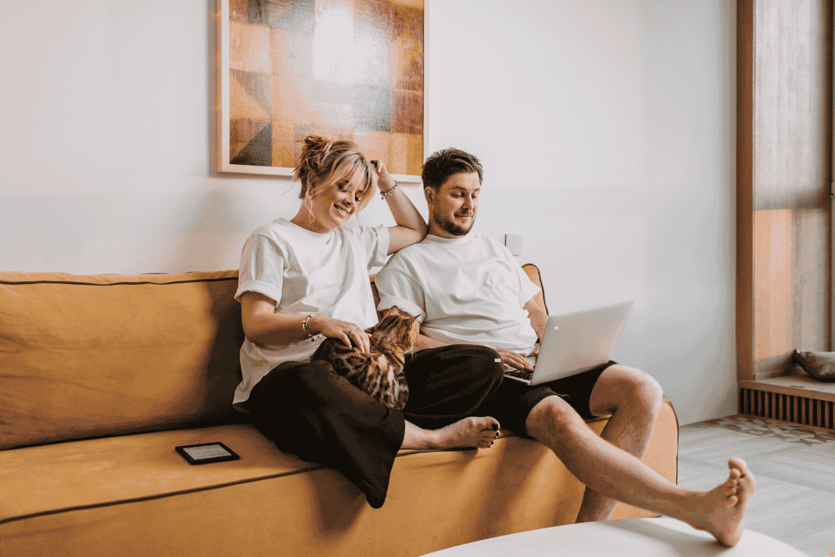 A couple sitting on a tan sofa at home in Albury Wodonga, smiling and relaxed with a cat on their lap, enjoying cool indoor comfort from an evaporative air conditioner on a warm summer day.
