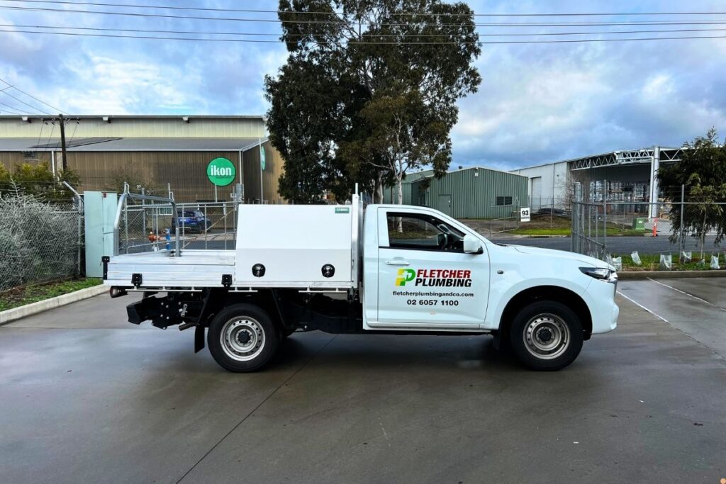 White Fletcher Plumbing & Co ute with tray back and toolboxes, parked at an industrial site